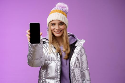 Lifestyle. Friendly cheerful confident blond girl in silver stylish winter jacket hat extend arm showing smartphone display advertising awesome new device app smiling self-assured recommend use mobile phone.