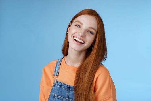 Positive outgoing lively redhead girl laughing joyfully having fun talking friendly friends tilting head chuckling joking funny life moments standing positive lucky blue background orange t-shirt. Lifestyle.