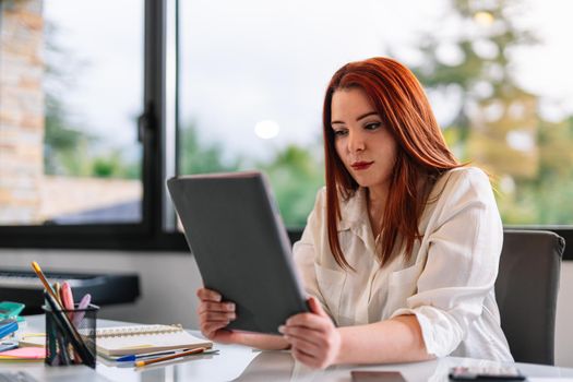 Portrait of a pretty young attractive girl with red hair in a white shirt using a tablet while teleworking at home. Enterprising young entrepreneur and self-employed girl working from home with tablet. White screen of tablet to paste any content. Large window in background with natural light.