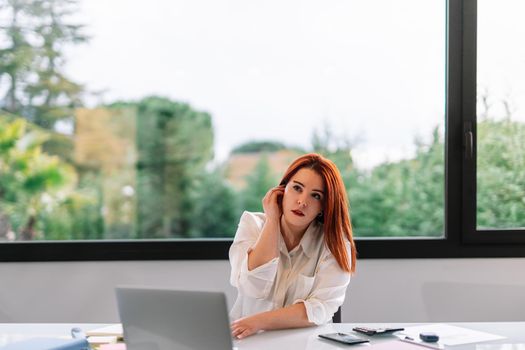 Pretty, beautiful young woman with red hair using her wireless headphones at home. Young entrepreneur and businesswoman teleworking at home in white shirt. Large window in background with natural light. She is using laptop and various office supplies.