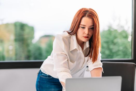 Attractive pretty young red-haired woman in white shirt and blue jeans standing looking at information on laptop while teleworking at home. Self-employed entrepreneur working from home on laptop. Large window in background with natural light.