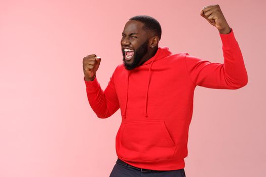 Lifestyle. Extremely happy bearded young 25s black guy triumphing happily yelling yeah raising clenched fists celebrating success winning bet lottery standing happily pink background cheer receive great news.