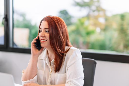 Pretty young red-haired woman looking at her smartphone while teleworking at home. Self-employed entrepreneur and businesswoman working from home or in an office while using her mobile. Large window in background with natural light.