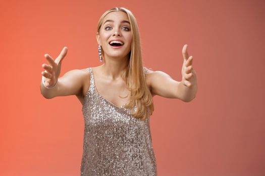 Excited charmign touched hearwarming young blond woman in silver party glamour dress stretch hands towards camera amused wanna hug cuddle hold cute puppy arms, standing red background.
