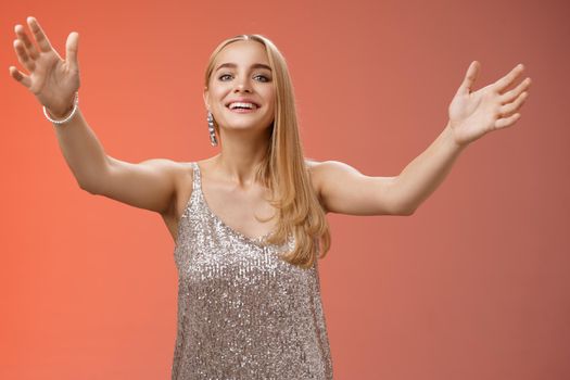Friendly charming tender hospitable attractive birthday girl in luxury silver dress welcoming guests throw party extend arms cuddle greeting people hug give cuddle smiling broadly, red background.