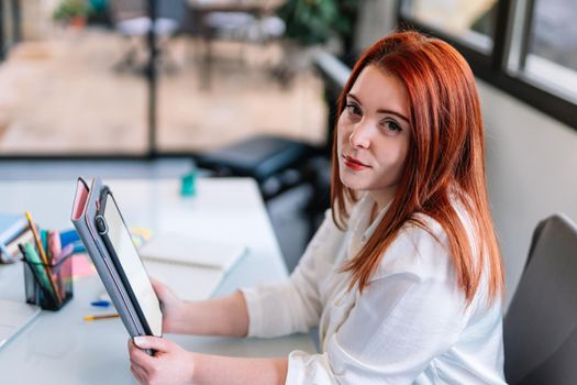 Portrait of a pretty young attractive girl with red hair in a white shirt using a tablet while teleworking at home. Enterprising young entrepreneur and self-employed girl working from home with tablet. White screen of tablet to paste any content. Large window in background with natural light.