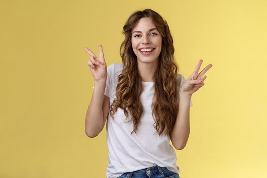 Friendly peaceful charming happy young girl curly long hair show peace victory signs having fun posing outgoing lively look camera amused laughing smiling broadly stand yellow background. Lifestyle.