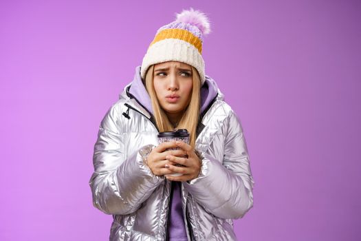 Trembling uncomfortable cute pouting young blond girl feel freezing cold winter snowy weather outside shaking low temperature hold take-away hot coffee cup warming up, purple background.