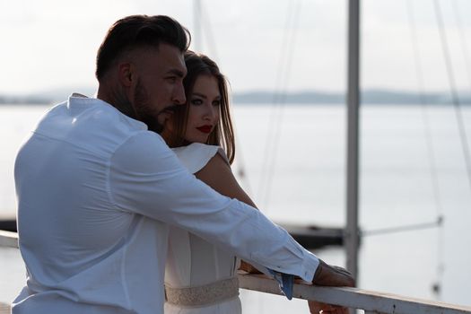 Loving couple in trendy outfits embracing while standing in port with boats in summer