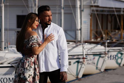 Loving couple in trendy outfits embracing while standing in port with boats in summer