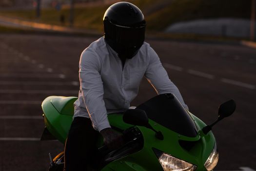 Unrecognizable stylish male motorcyclist in helmet sitting on parked motorbike in city on background of sunset sky