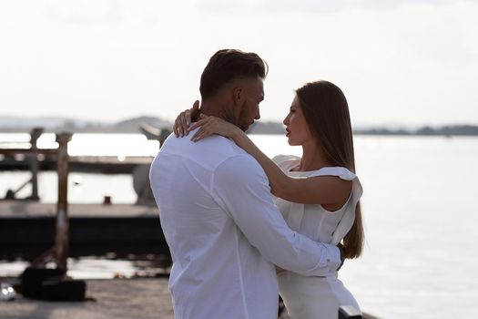 Loving couple in trendy outfits embracing while standing in port with boats in summer