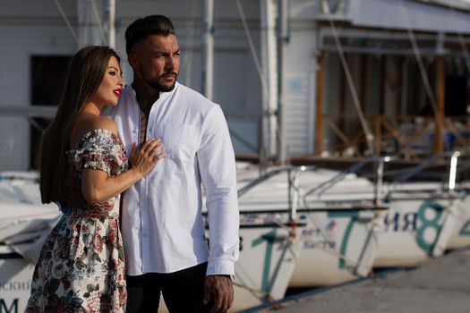 Loving couple in trendy outfits embracing while standing in port with boats in summer