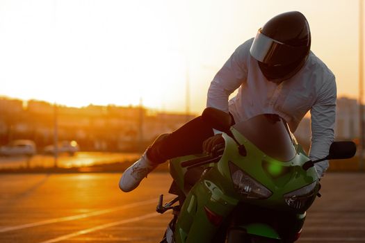 Unrecognizable stylish male motorcyclist in helmet sitting on parked motorbike in city on background of sunset sky