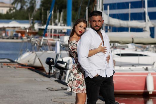 Loving couple in trendy outfits embracing while standing in port with boats in summer