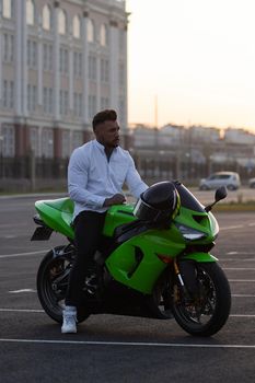 Side view of handsome male motorcyclist sitting on modern cool motorbike parked on asphalt parking against sunset sky