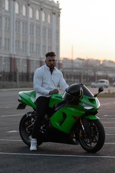 Side view of handsome male motorcyclist sitting on modern cool motorbike parked on asphalt parking against sunset sky