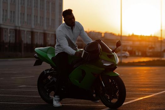 Side view of handsome male motorcyclist sitting on modern cool motorbike parked on asphalt parking against sunset sky