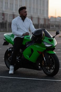 Side view of handsome male motorcyclist sitting on modern cool motorbike parked on asphalt parking against sunset sky