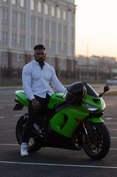 Side view of handsome male motorcyclist sitting on modern cool motorbike parked on asphalt parking against sunset sky