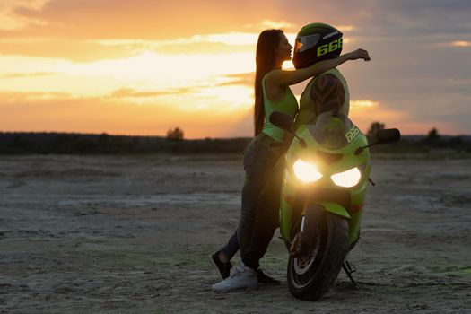 Side view of couple in love tenderly hugging near motorcycle parked on sandy seashore against sundown sky