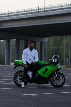 Side view of handsome male motorcyclist sitting on modern cool motorbike parked on asphalt parking against sunset sky