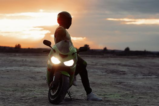 Side view of stylish male motorcyclist in helmet standing near modern motorbike on sandy seashore in evening