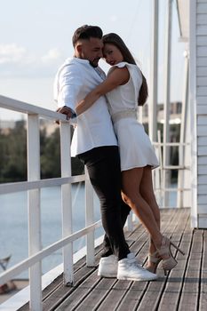 Loving couple in trendy outfits embracing while standing in port with boats in summer