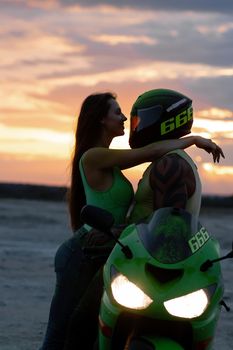 Side view of couple in love tenderly hugging near motorcycle parked on sandy seashore against sundown sky