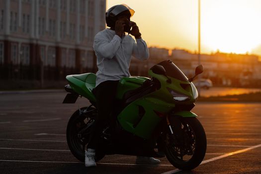 Unrecognizable stylish male motorcyclist in helmet sitting on parked motorbike in city on background of sunset sky