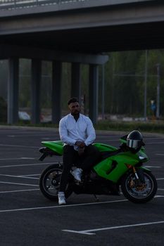 Side view of handsome male motorcyclist sitting on modern cool motorbike parked on asphalt parking against sunset sky