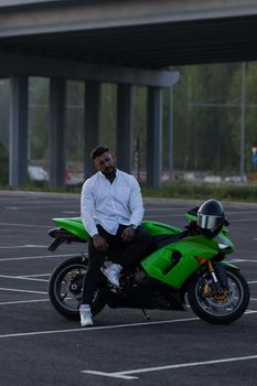 Side view of handsome male motorcyclist sitting on modern cool motorbike parked on asphalt parking against sunset sky