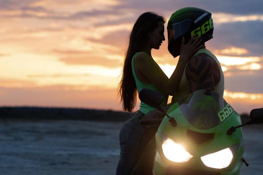 Side view of couple in love tenderly hugging near motorcycle parked on sandy seashore against sundown sky