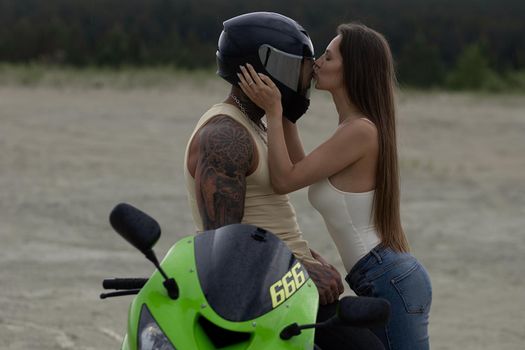 Side view of stylish loving couple cuddling near motorbike on beach at sunset
