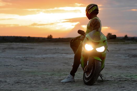 Side view of stylish male motorcyclist in helmet standing near modern motorbike on sandy seashore in evening