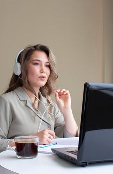 beautiful asian girl is sitting with headphone at a computer and saying something. there are a cup of coffee, a copybook and a pencil on a table