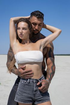 Seductive female in swimsuit and fit male with naked torso hugging while standing on sandy seashore on sunny day in summer