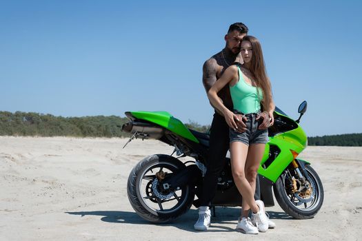 Serious male with tattoos and beard standing and embracing sensual girlfriend with long brown hair against green motorbike in sandy field in nature in sunny day