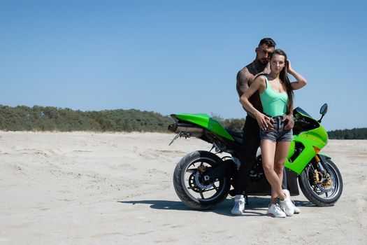 Serious male with tattoos and beard standing and embracing sensual girlfriend with long brown hair against green motorbike in sandy field in nature in sunny day