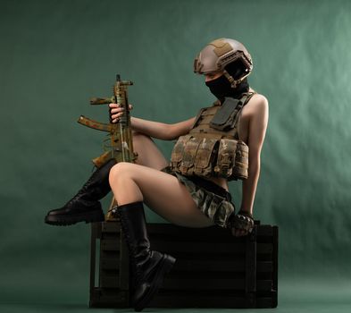 girl soldier armed with an automatic rifle in military clothes sits on a gun case on a green background