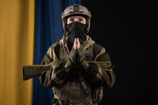 male soldier in a military uniform and helmet with a weapon on the background of the national flag of Ukraine prays for peace