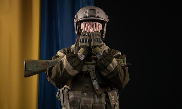 a male soldier in a military uniform and helmet with a weapon on the background of the national flag of Ukraine covered his eyes with his hands