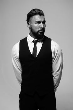 Bearded Hispanic guy in dark vest and white shirt with tie looking at camera with hands in pockets in studio