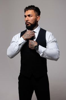 Confident Hispanic bearded man with tattooed hands in white shirt and black trousers adjusting tie and looking away while standing against gray background