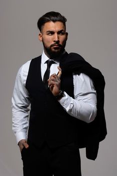 Bearded Hispanic guy in dark vest and white shirt with tie looking at camera with hands in pockets in studio