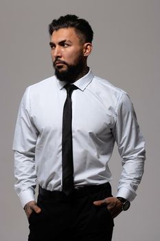 Bearded Hispanic guy in dark vest and white shirt with tie looking at camera with hands in pockets in studio