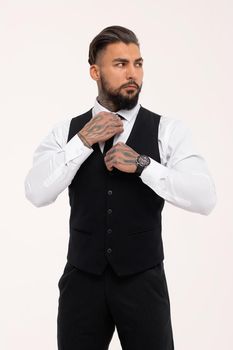 Confident Hispanic bearded man with tattooed hands in white shirt and black trousers adjusting tie and looking away while standing against gray background