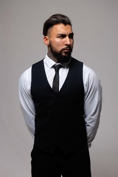 Bearded Hispanic guy in dark vest and white shirt with tie looking at camera with hands in pockets in studio