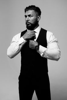 Confident Hispanic bearded man with tattooed hands in white shirt and black trousers adjusting tie and looking away while standing against gray background
