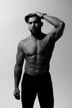 Hispanic shirtless male model with muscular tattooed torso standing with hands in pockets and looking away on gray backdrop